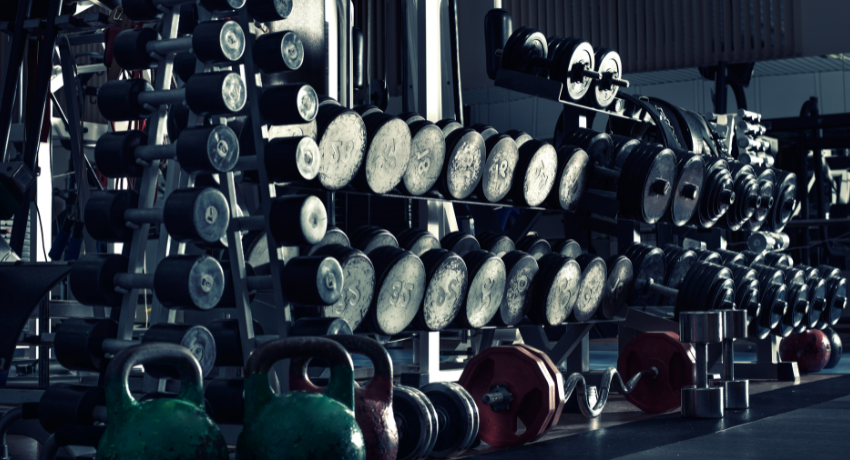 Conjunto de halteres, kettlebells e barras organizados em suportes dentro de uma academia com equipamentos de musculação.